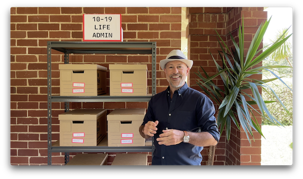 Johnny standing in front of a shelving unit, with four storage boxes visible. He's using it as the analogy to the system's 'areas' and 'categories'.